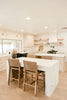 A kitchen with white marble islands, woven bar stools, and light wood flooring